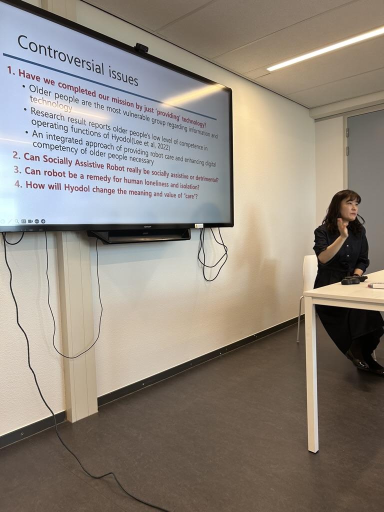 Sochung lee presenting in the classroom. She is sitting in a chair behind a white desk and in front of a smart white board. 