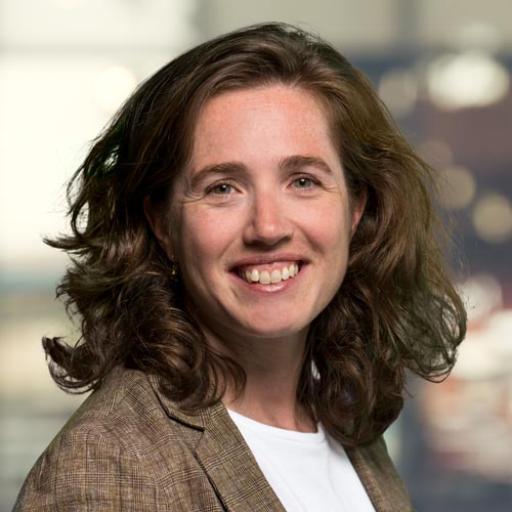 A picture of Anne Fleur van Veenstra. A woman with dark hair standing in front of a blurred background, wearing a white t-shirt with a light brown checkered jacket. 