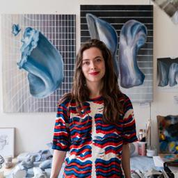 Gill Baldwin standing in her studio. She has dark hair and is smiling. She is wearing a red-white and blue blouse.