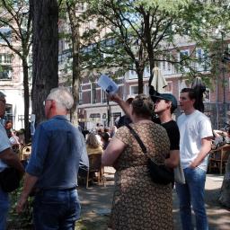 Group of people in discussion, one of them is pointing upwards. They are standing outside on a square. 