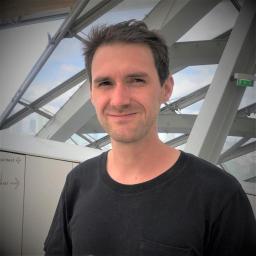 A picture of Vivien, a man with dark hair wearing a black t-shirt. He is standing in front of a iron sculpture. 